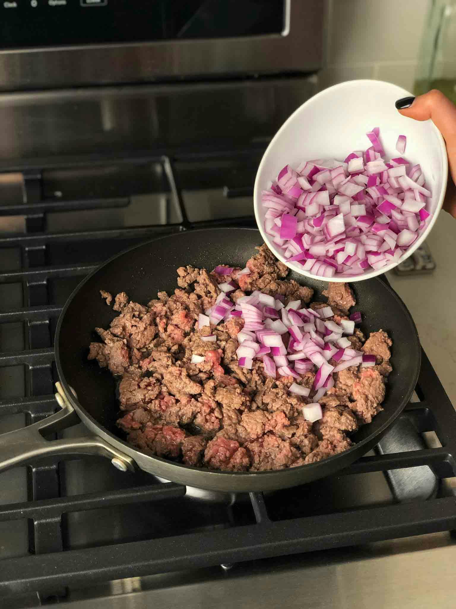 Ground beef cooking in skillet with onions being added.