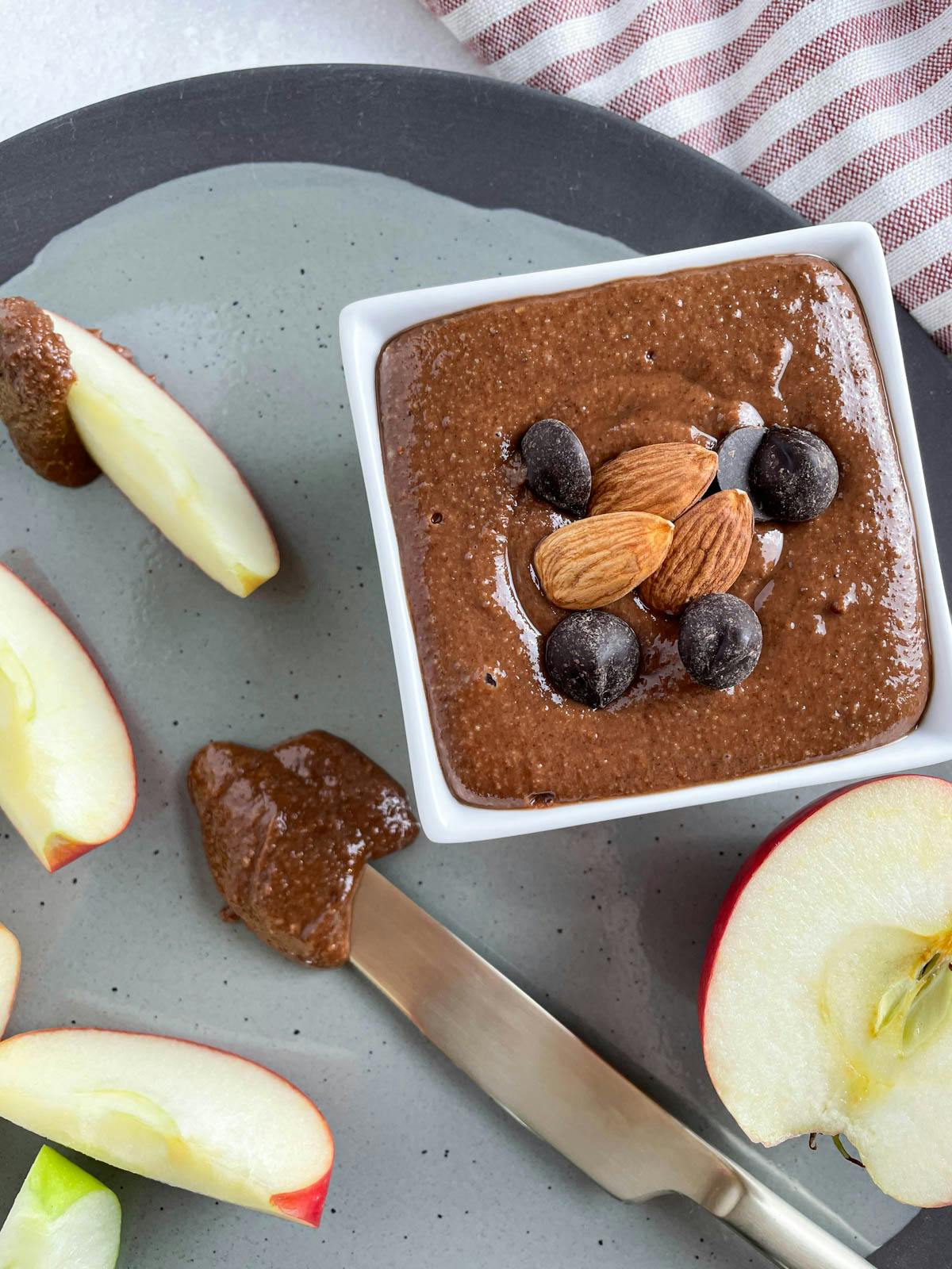 A small white dish of chocolate almond butter by some sliced apples.
