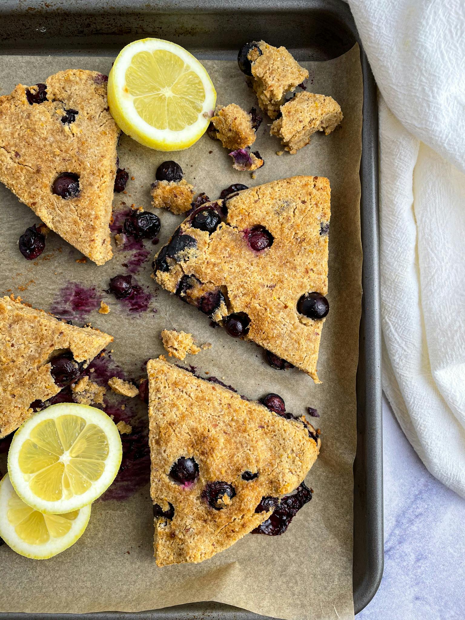 Gluten-free blueberry lemon scones spread out on a baking sheet with slices of lemon around them. There are crumbles of blueberry lemon scones on the top of the baking sheet.