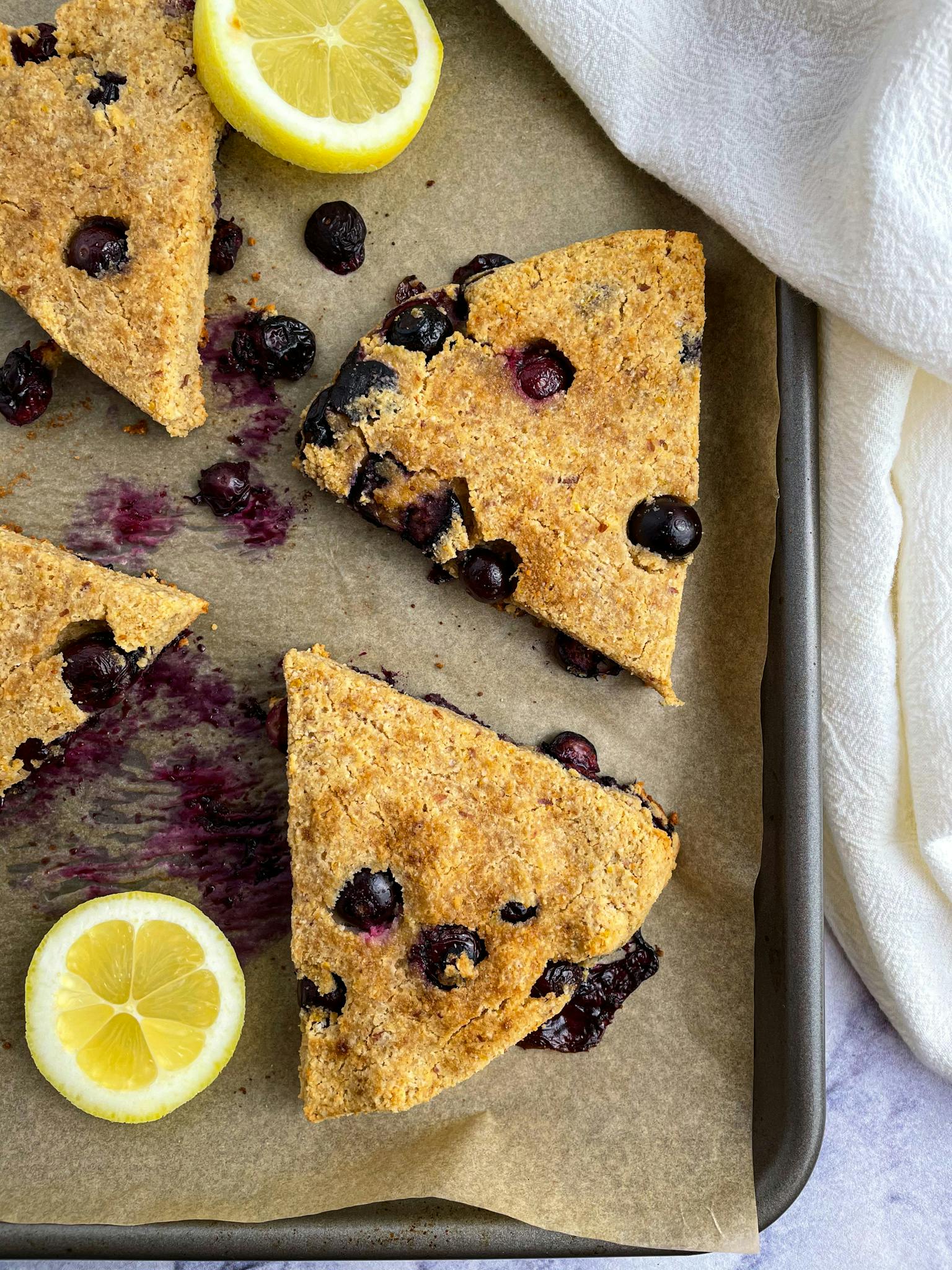 Gluten-free blueberry lemon scones spread out on a baking sheet with slices of lemon around them.